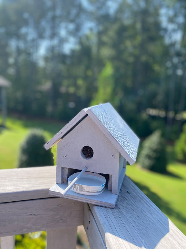 Gray 3D-printed birdhouse with a round entrance hole and a feeding tray, mounted on a deck railing