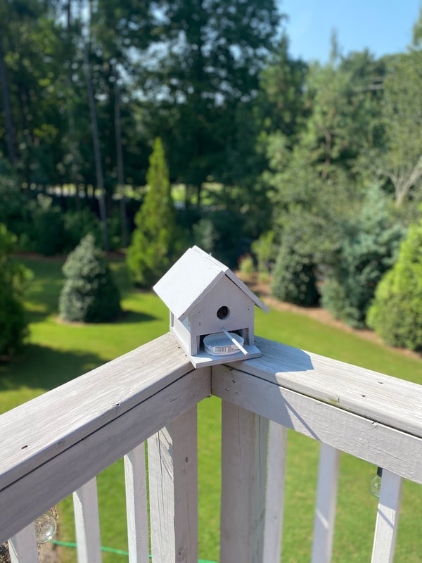 The 3D-printed birdhouse feeder on a deck railing with trees behind it
