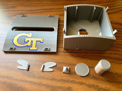 3D-printed gray box and lid with a gold and navy Georgia Tech logo, laid out with small printed parts on a wooden table